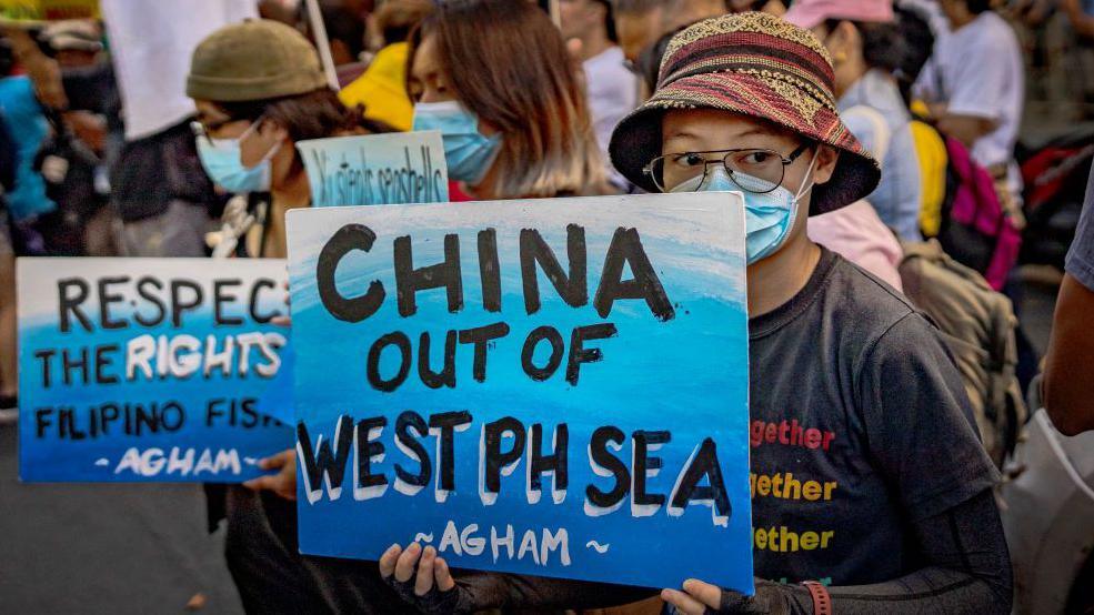 A proester in Manila, wearing a hat, eyeglasses and face mask carrying a blue sign with the words China out of West Philippine Sea in bold black font