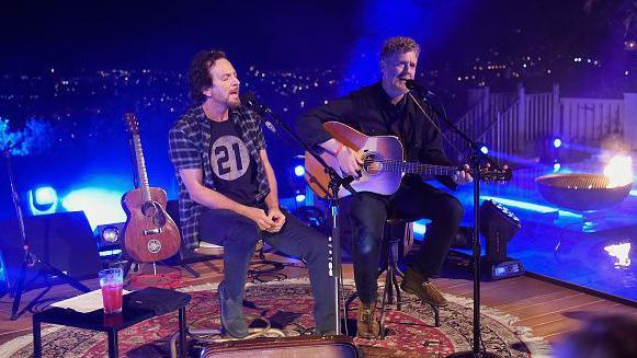 Eddie Vedder is wearing a checked shirt and black t-shirt and jeans and has dark hair and a goatee beard he is singing beside Glen Hansard who is playing guitar and singing.