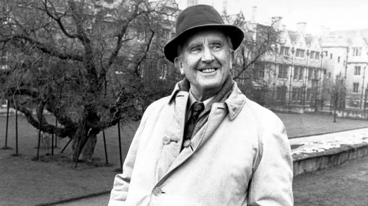 A black and white image of JRR Tolkien smiling for a photo while on a walk outside. A row of houses could be seen behind him.