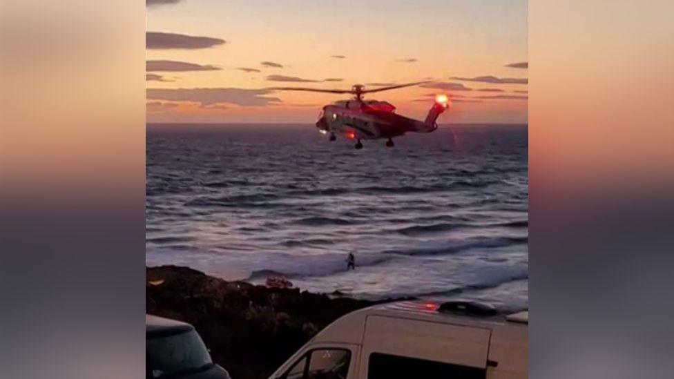 The image shows a coastguard rescue helicopter hovering low over the sea close to a shoreline at dusk. In the foreground there are two parked vans beside a coastal road or car park, while waves are breaking near the shore below and a small figure can be seen in the water or surf; the helicopter's red lights are illuminated against a sunset sky, suggesting an ongoing maritime rescue or emergency response operation near a beach.