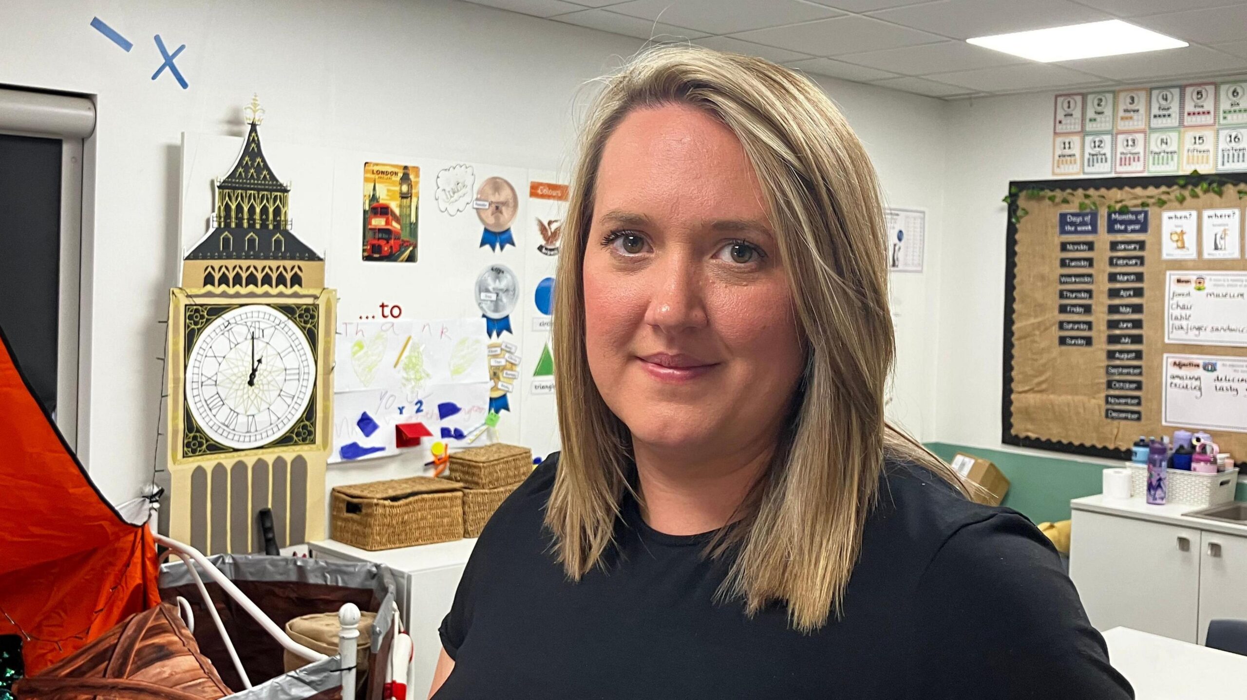 A head and shoulder shot of Helen Blythe. She has shoulder length straight, blonde hair and is wearing a black T-shirt. She is stood in a school classroom which has walls decorated with posters.