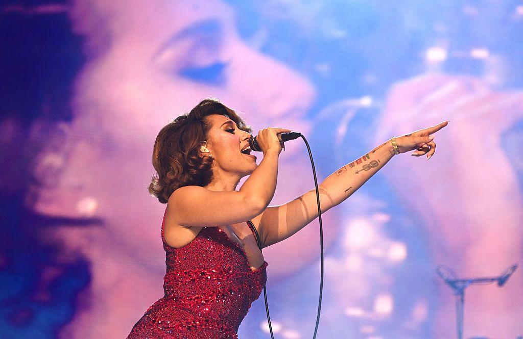 Raye performs on stage during her 'This Tour May Contain New Music' tour at The O2 Arena on May 19, 2026 in London, England. She is wearing a red, sparkly gown and is singing into a wired microphone. The backdrop is an enlarged video screen