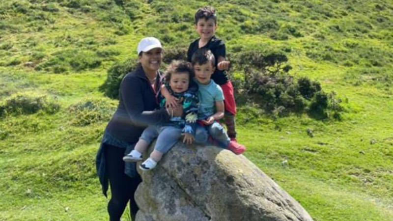 A woman is seen standing in a field with three boys sat on a rock.