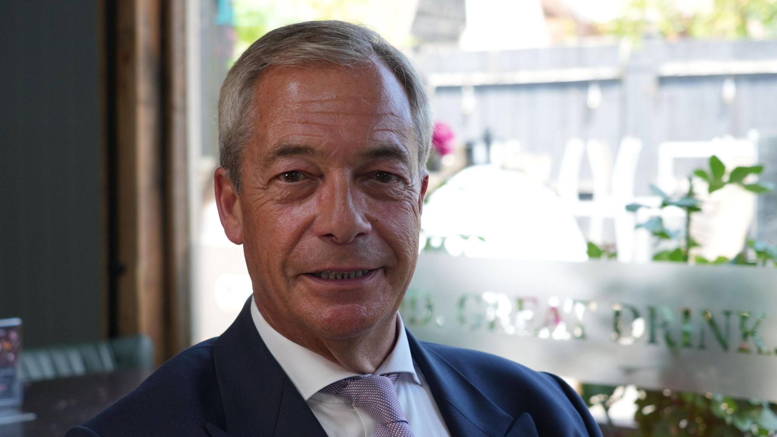 Nigel Farage smiles for the camera. He has short grey hair and is wearing a blue jacket with a white shirt and purple tie.