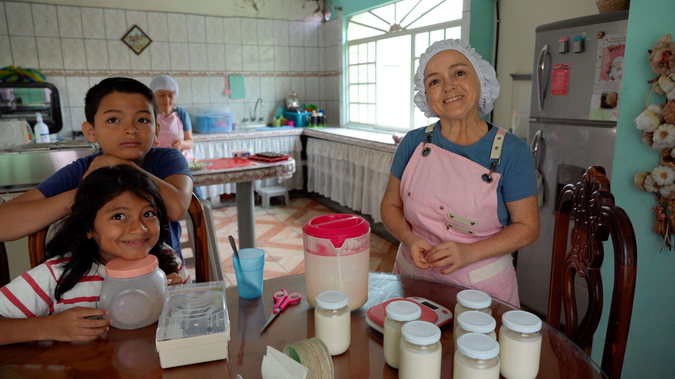 María Luisa, pictured with her and her sister's children. She is at the right of the frame, smiling, with a pink apron and hair net. The little girl smiles at the camera while sitting on a chair, she has long dark hair, while her cousin leans on the back of the chair. He has short dark hair. María del Cisne is working in the background.