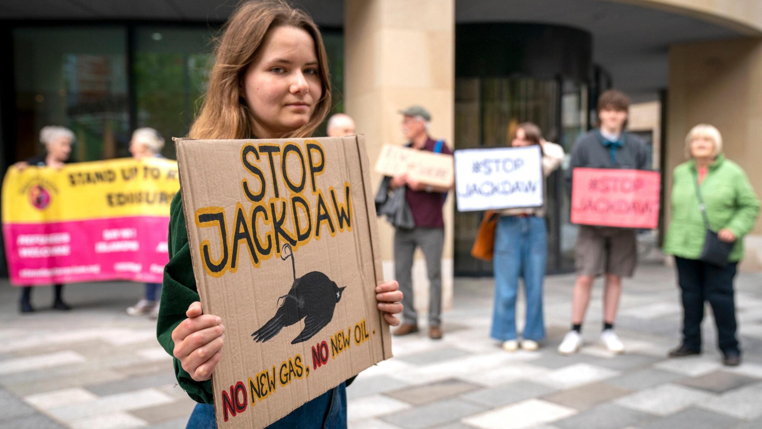 Environmental campaigners from Friends of the Earth protest outside a UK government building in Edinburgh against the decision to approve Shell's Jackdaw gas field in June 2022. A woman with shoulder-length brown hair is in the foreground holding as placard reading: Stop Jackdaw. No new gas, no new oil. Begins her is a row of campaigners holding placards saying #Stop Jackdaw.