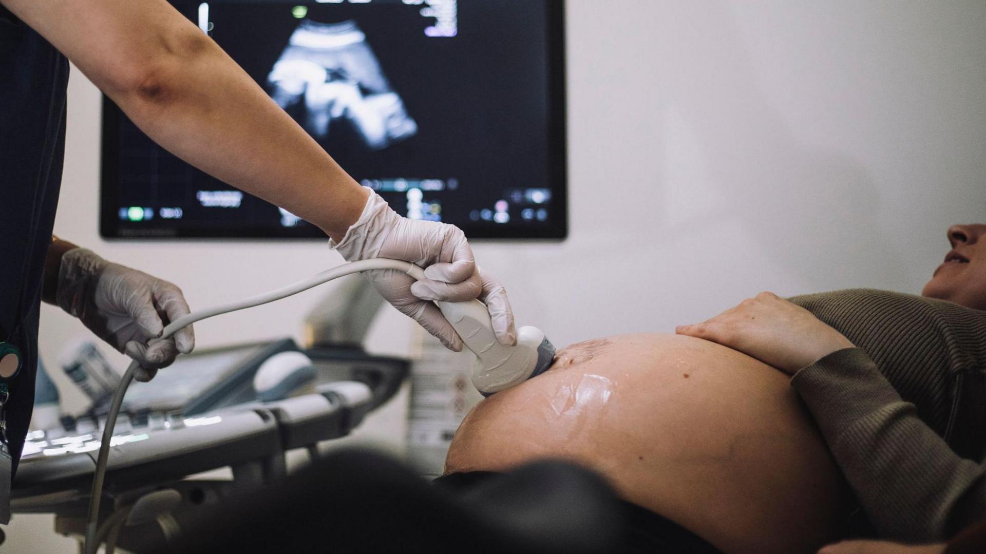 A healthcare professional wearing gloves performs an ultrasound examination on a pregnant woman. The ultrasound probe is placed on the patient's abdomen, while a monitor in the background displays the ultrasound image. The scene takes place in a clinical medical setting, with diagnostic equipment visible beside the examination table.