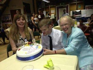 Charlie, his sister and mum, sit around a decorated birthday cake with candles, while other guests are visible in the background