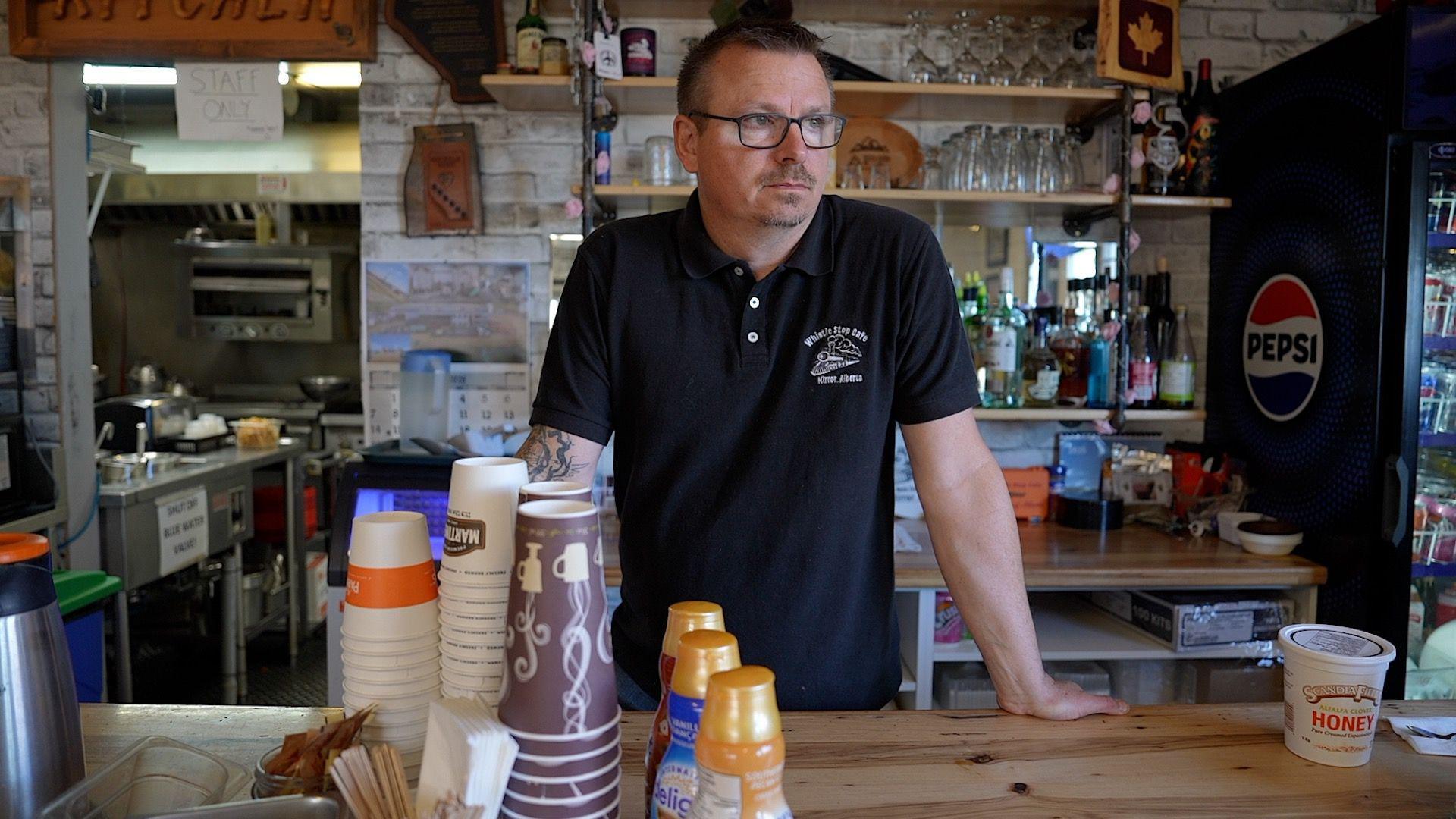 Chris Scott stands behind the counter at his Whistle Stop cafe. He is wearing a collared black t-shirt with his cafe's logo in white. In front of him are disposable coffee cups and three bottles of coffee creamer. Behind him are bar shelves with alcohol bottles and drinking glasses. He is wearing square-framed seeing glasses, has a moustache and goatee and a short haircut, and is looking off to the right.