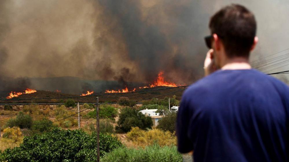 Flames and black smoke are seen rising along the horizon from a hilly area in the background, while a man in the foreground looks towards it with his hand to his mouth