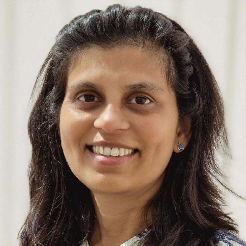 Priya Kapadia with long black hair pushed back off her face with a headband, smiling at the camera