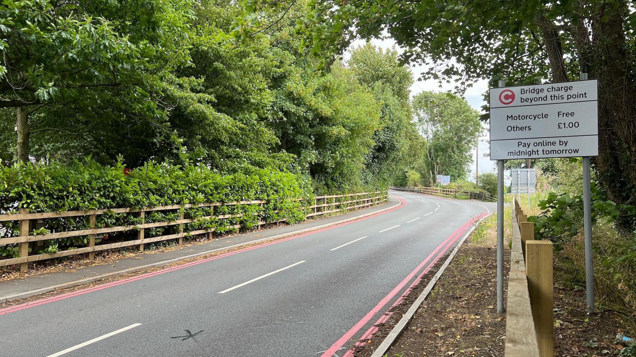 A view of the road up to the Warburton toll bridge. On the right hand side of the image there is a sign that says "bridge charge beyond this point" and it says that is free for motorcycles and £1 for other vehicles. It also says "pay online by midnight tomorrow night".