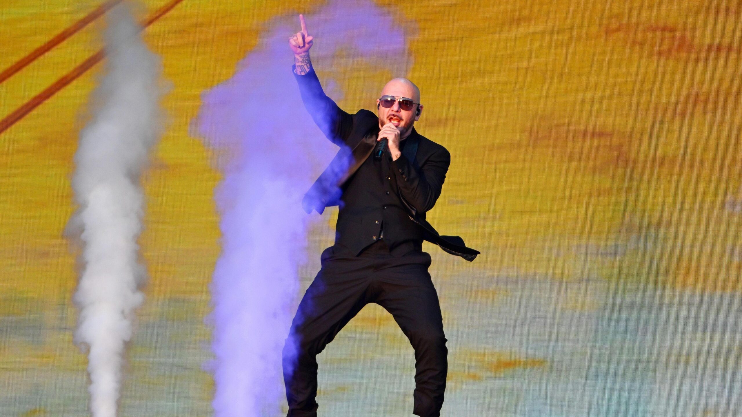 Pitbull performs on stage as smoke machines go off in the background. He wears a black suit and sunglasses.
