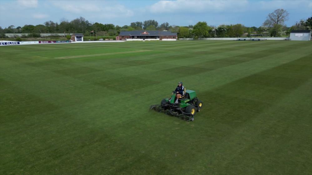 Jimmy Perchard on his lawn mower at Farmers field ground with the club house in the background.