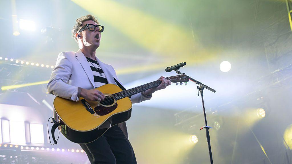 Callum Beattie is playing guitar on stage. The picture has been taken from below, looking up at him. He is wearing thick-rimmed, black glasses, a black and white striped T-shirt, jeans and a white suit jacket. There are yellow and white stage lights illuminating the picture