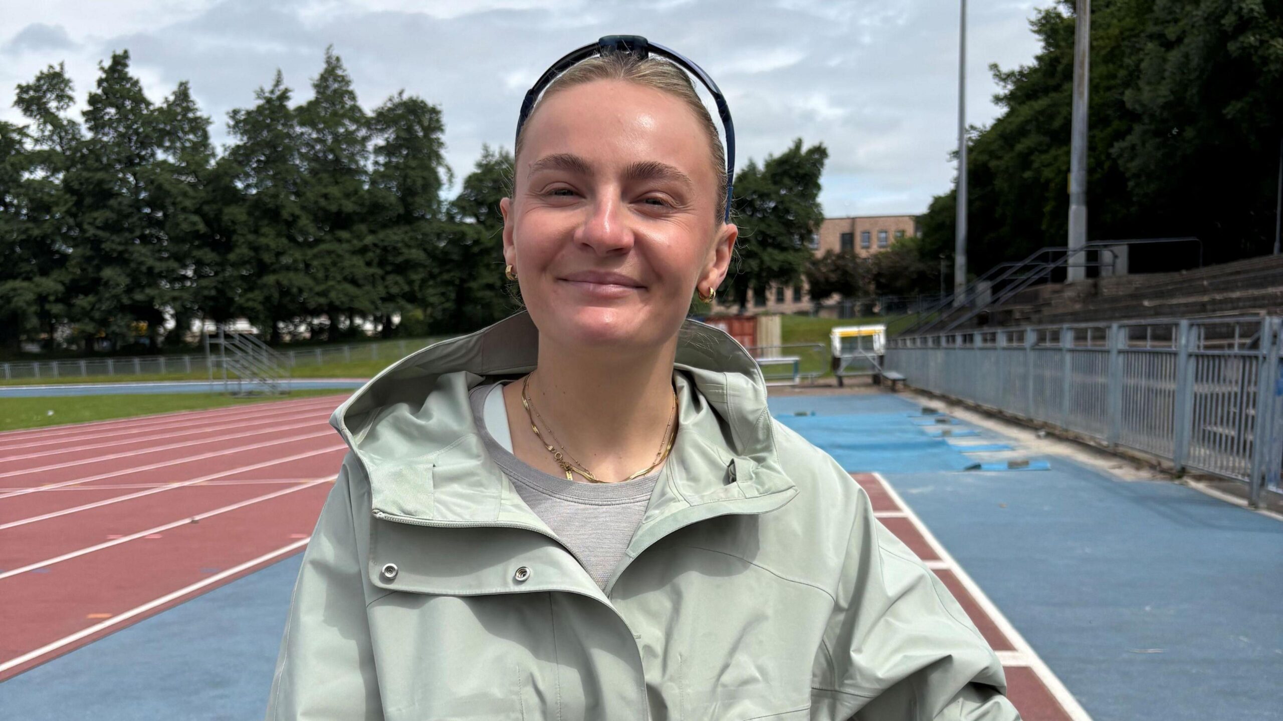 Head and shoulders image of wheelchair racer Melanie Woods. She is on running track and is facing into the camera. She is white and has brown hair tied back and black sunglasses on the top of her head. She's wearing a light khaki green rain jacket and a grey t-shirt and has a gold necklace and gold earrings
