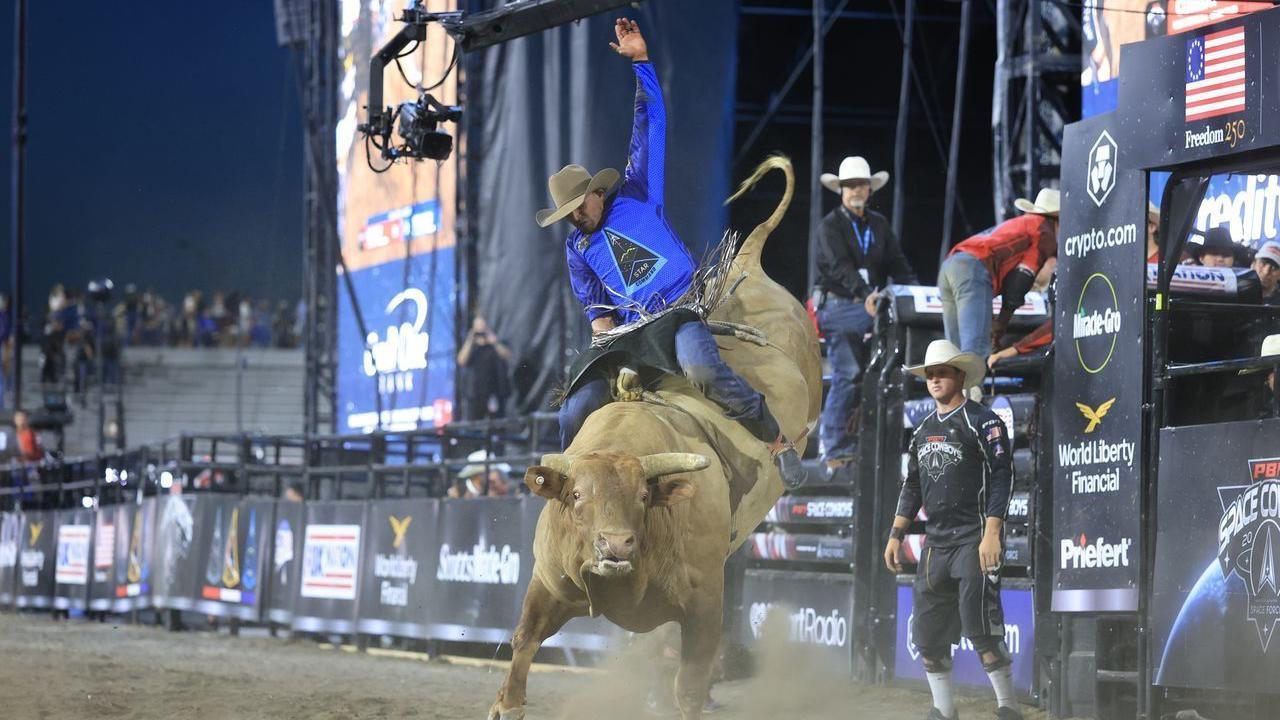 A professional bull rider, wearing a cowboy hat and blue shirt featuring the triangular logo of US Space Force, clings on while riding on the back of a massive, brown horned bull in a stadium filled with dirt