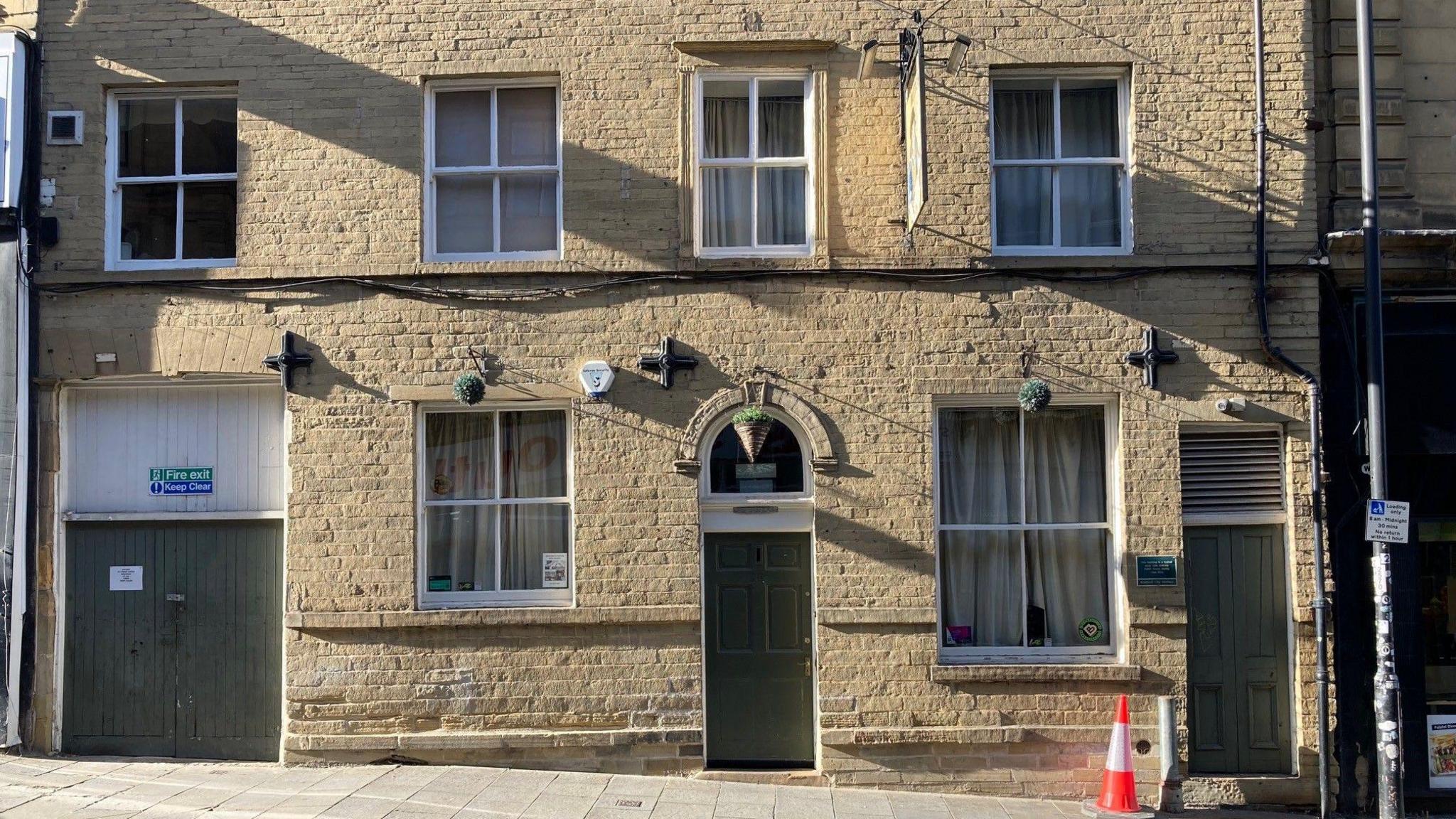 The sandy-coloured, Yorkshire stone front of the pub with cream curtains drawn in all its wood-framed windows which are painted white.