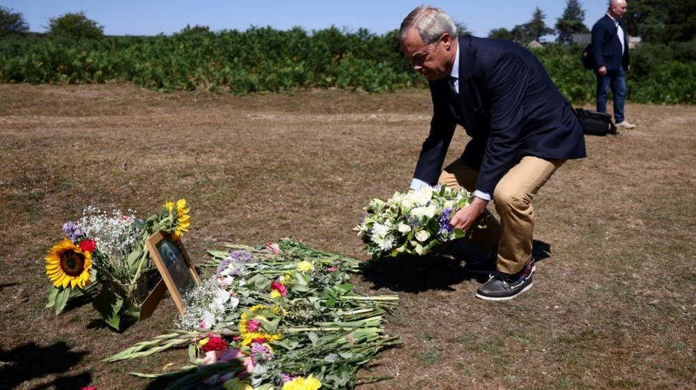 Nigel Farage, wearing a navy blazer, blue shirt, beige trousers and boat shoes is seen bending down to place a wreath of cream and lilac flowers. On the ground, bunches of flowers have already been laid, alongside a framed black and white photograph of Ann Widdecombe