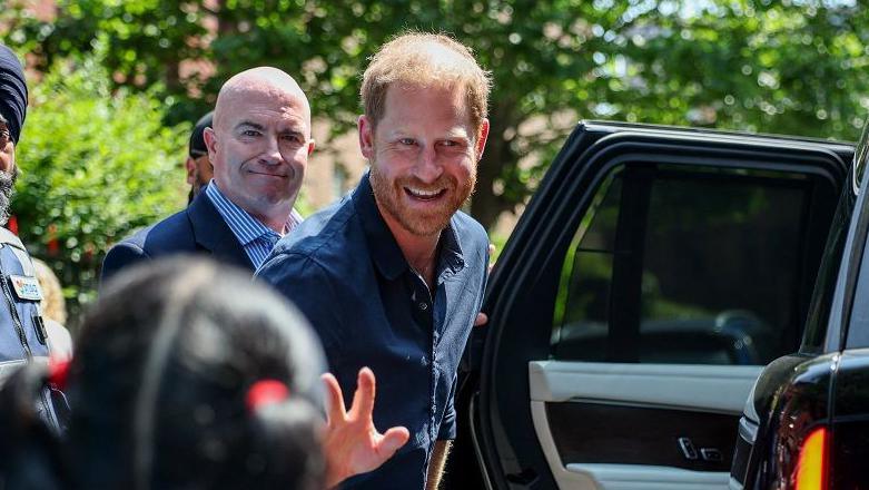 Britain's Prince Harry waves at a child and a crowd as he gets into his car after leaving a hospital visit. He is wearing a blue shirt, and is looking to the left of the camera and smiling