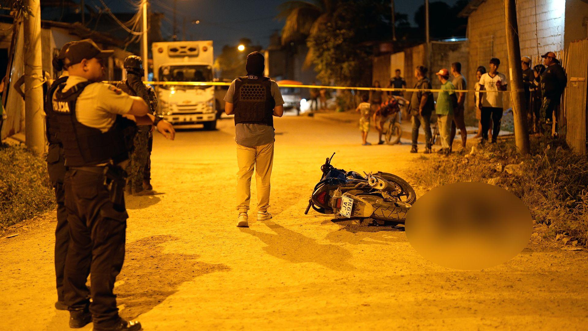 Police stand beside a corpse, lying next to a toppled motorcycle on the street. Onlookers watch from behind police tape.