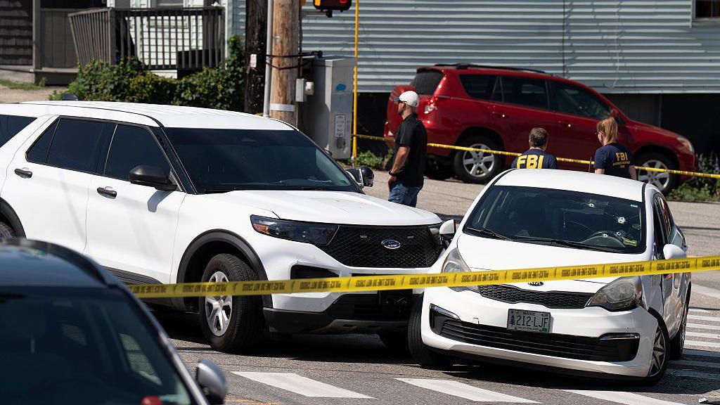 Two white cars that collided are behind yellow police tape. The windscreen of one car has bullet holes in it
