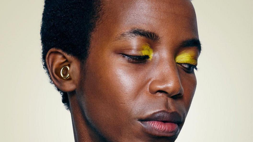 A close-up of someone wearing yellow eye shadow. In their ear is a gold loop-style ear-plug.