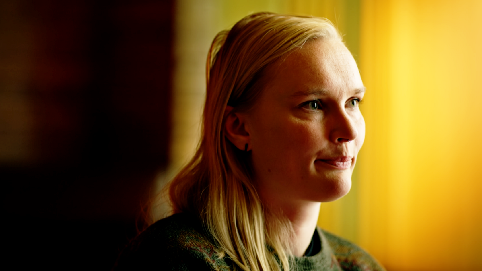 A close-up portrait of Alex looking serious, with pursed lips. Her face is turned to the side and she is looking towards the right of the frame. She has blonde hair and wears a dark top and small earring. The background is softly blurred in shades of yellow, gold, and dark brown.