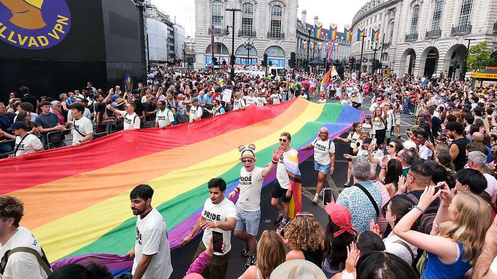 A large crowd of spectators lines the street as a group of parade participants carries a massive, elongated rainbow Pride flag down a central London road flanked by historic stone buildings.