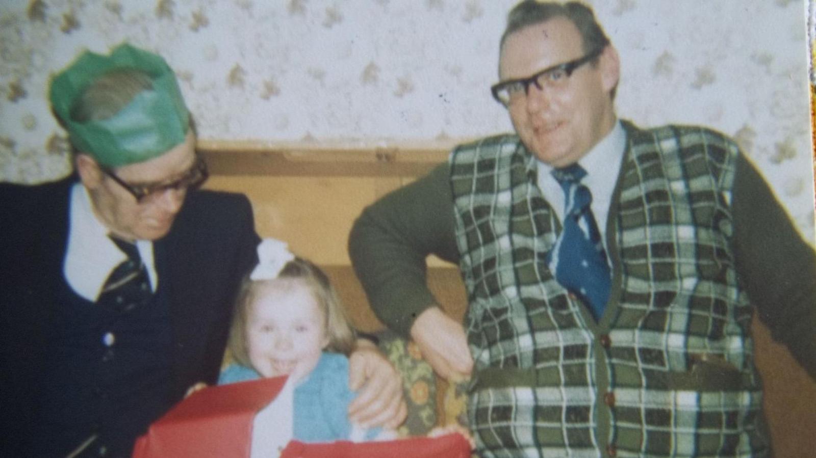 A colour family photograph from the 1970s. Anthony Littler, a large man in glasses and a green checked tank top with a blue tie, sits smiling on a sofa. Beside him to the left an older man in glasses and a green paper party hat leans towards a small girl in a blue dress opening a large present wrapped in red paper.