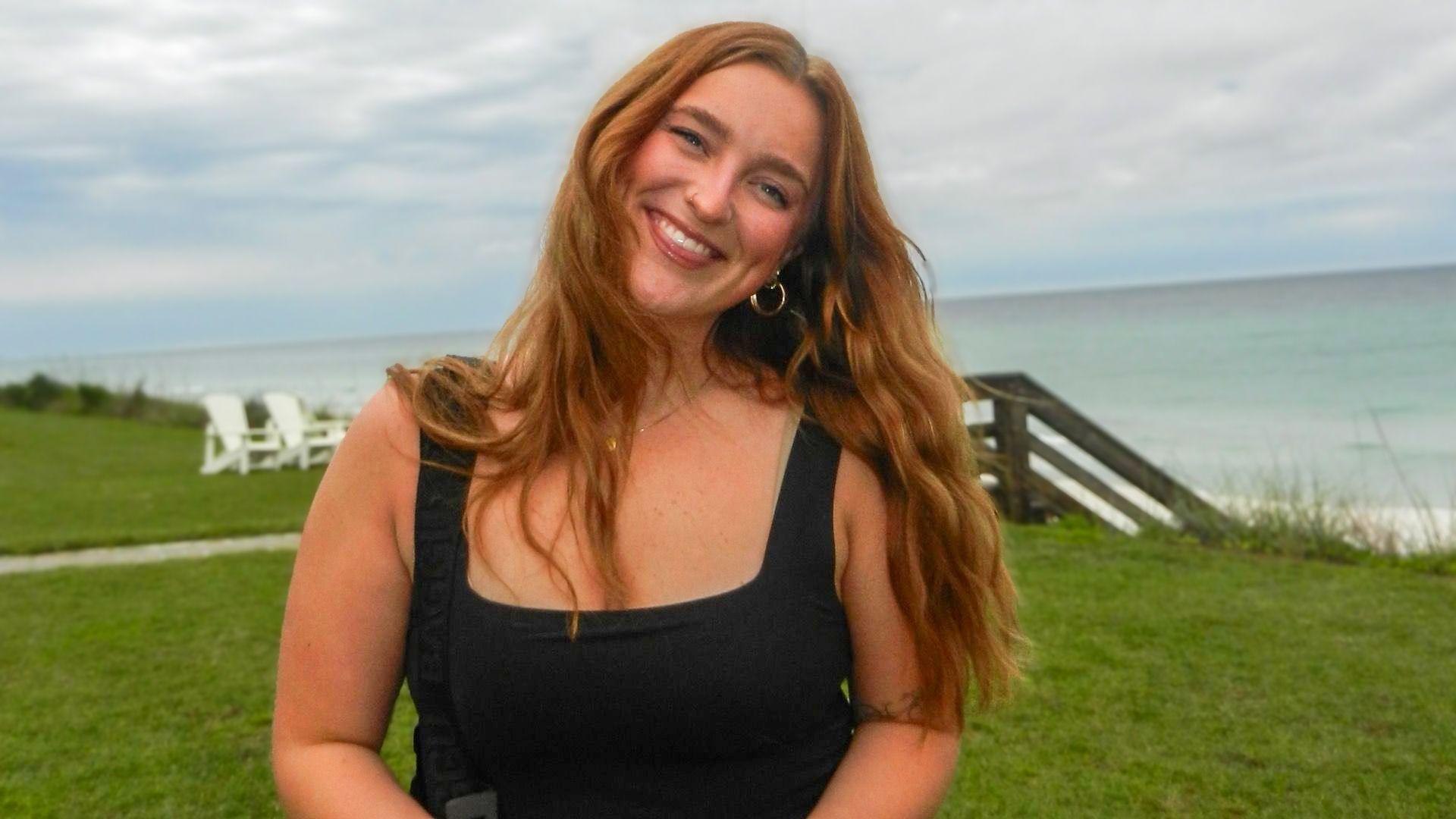 Lizzy has long reddish hair and is wearing a black top whilst standing on a grassy cliff near the beach. The sky is cloudy and wooden steps to the beach and white chairs are visible behind her.