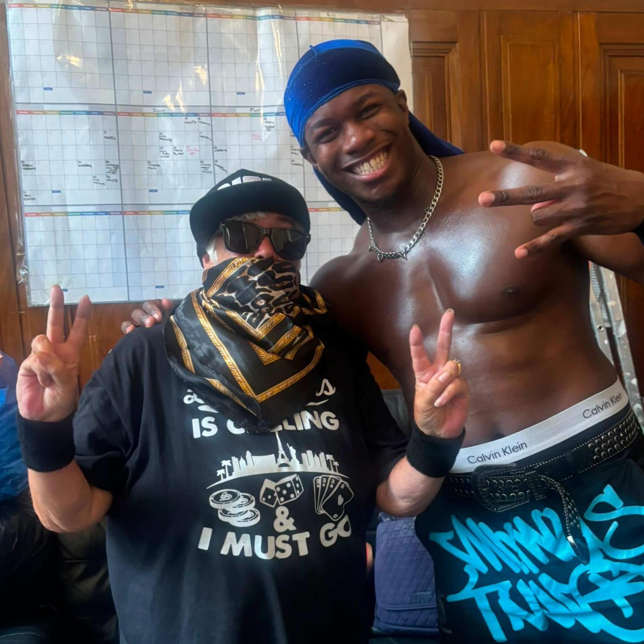 Susie Matthews, 73 and Man Like DeReiss photographed together during Cardiff wrestling showcase. Matthews is wearing a scarf covering the bottom half of her face, sunglasses and a black cap. She is doing a peace sign with both hands. DeReiss is stood next to her with his hand on her shoulder. He is wearing no t-shirt, a silver chain necklace and a blue durag cap.