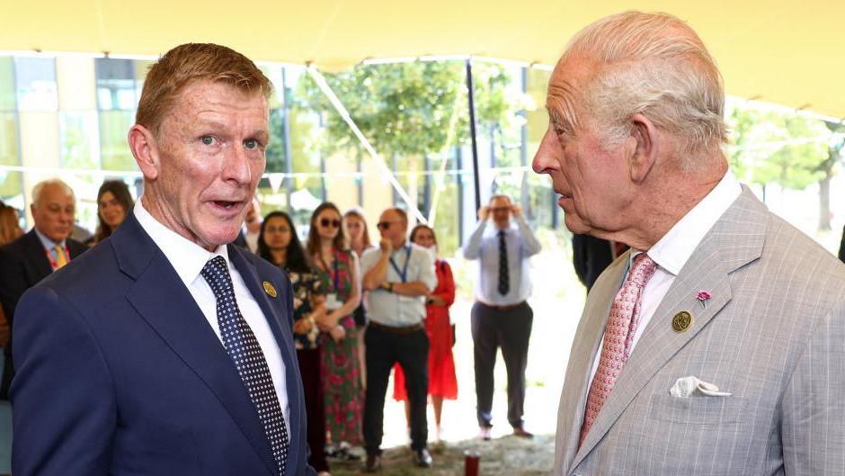 Tim Peake and King Charles stand next to each other talking. Both men are wearing suits