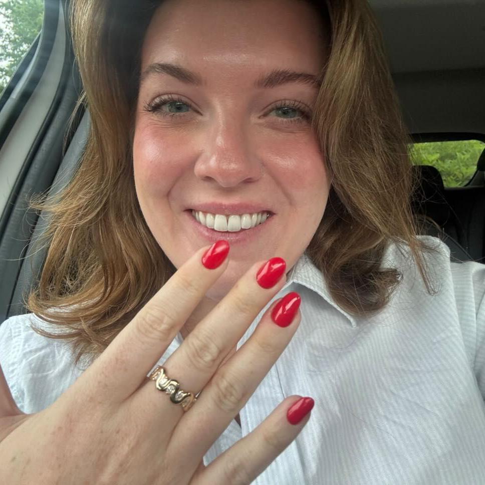 Alex Proie sitting in a car holding up her hand with a gold and diamond wave design ring on her middle finger and smiling