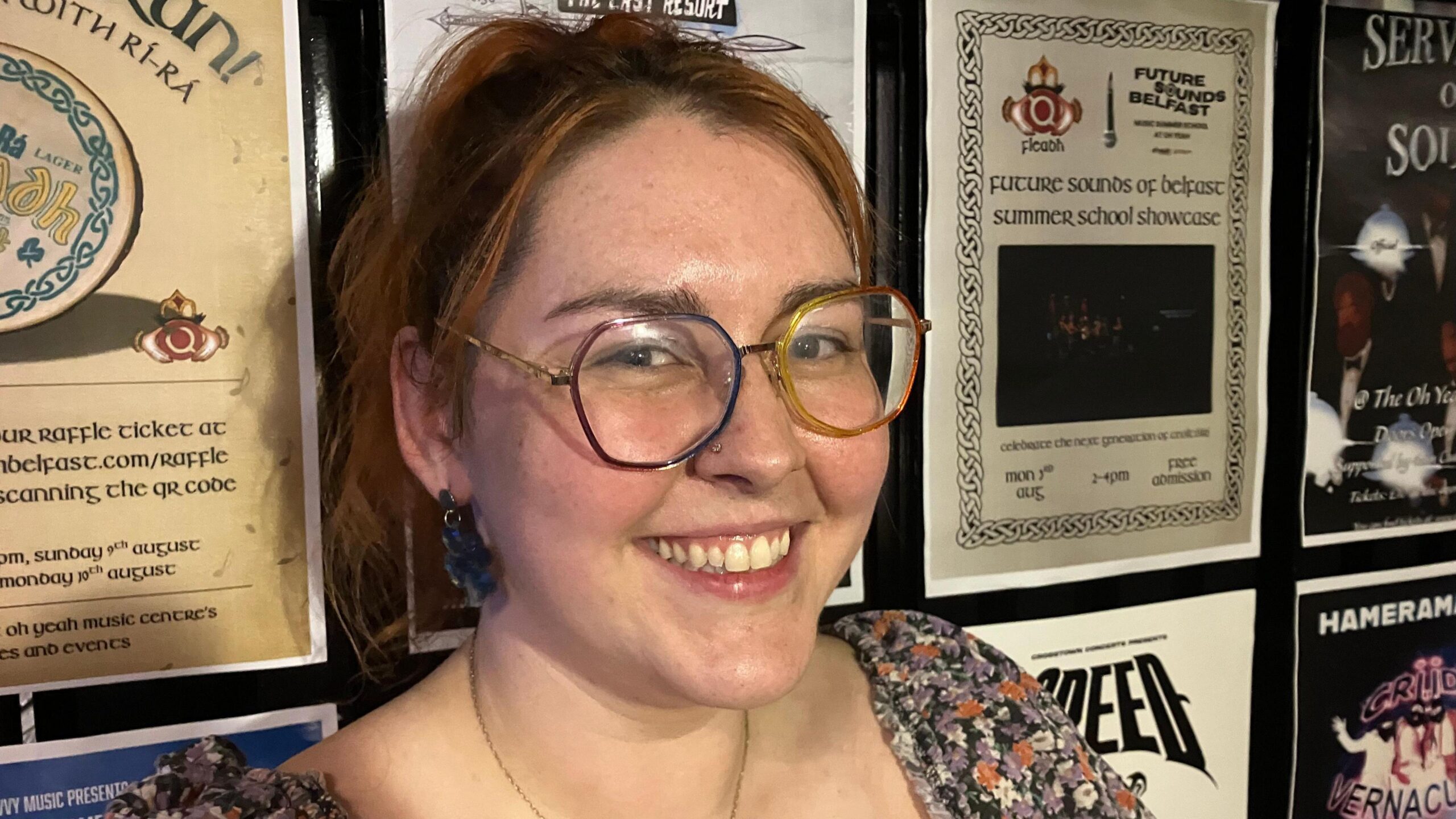 A woman with dark red hair, tied up, smiling at the camera. She is wearing glasses and a floral dress.