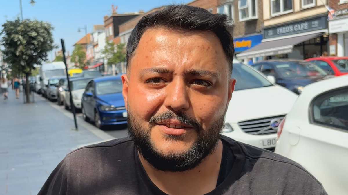 Memmzy Ozy has short black hair and a beard. He is wearing a black T-shirt in front of a row of parked cars.