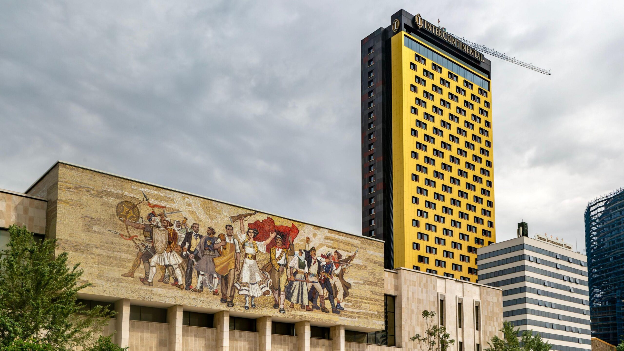 Tirana's central Skenderbeg Square. In the foreground is a large mural from Albania's communist era in the socialist-realist style. Behind this building there are cranes and skyscrapers under construction.