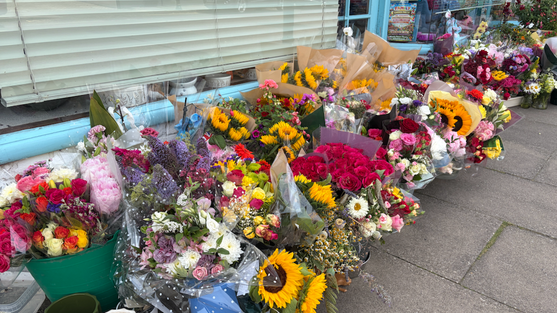Dozens of bouqets of flowers line the pavement outside a shop in buckets and vases. They include sunflowers, roses and other brightly-coloured arrangements.