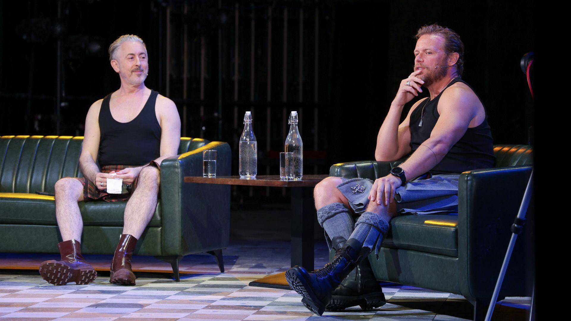 Two speakers sit on green leather armchairs on a stage during a live discussion. A small table between them holds two glass bottles of water and two drinking glasses. Both speakers wear black sleeveless tops with kilt-style skirts and boots, and one speaker is wearing a headset microphone while speaking. The stage is lit with blue and purple lighting against a dark background.