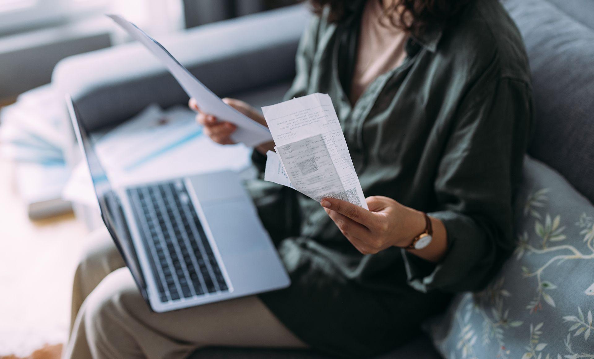 Woman overwhelmed with bills to pay, she is sitting on her sofa with a laptop holding receipts
