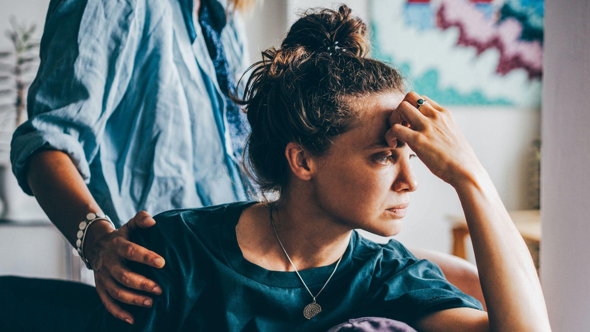 A sad woman sitting on a sofa, a person behind them has placed their hand on her shoulder in empathy.