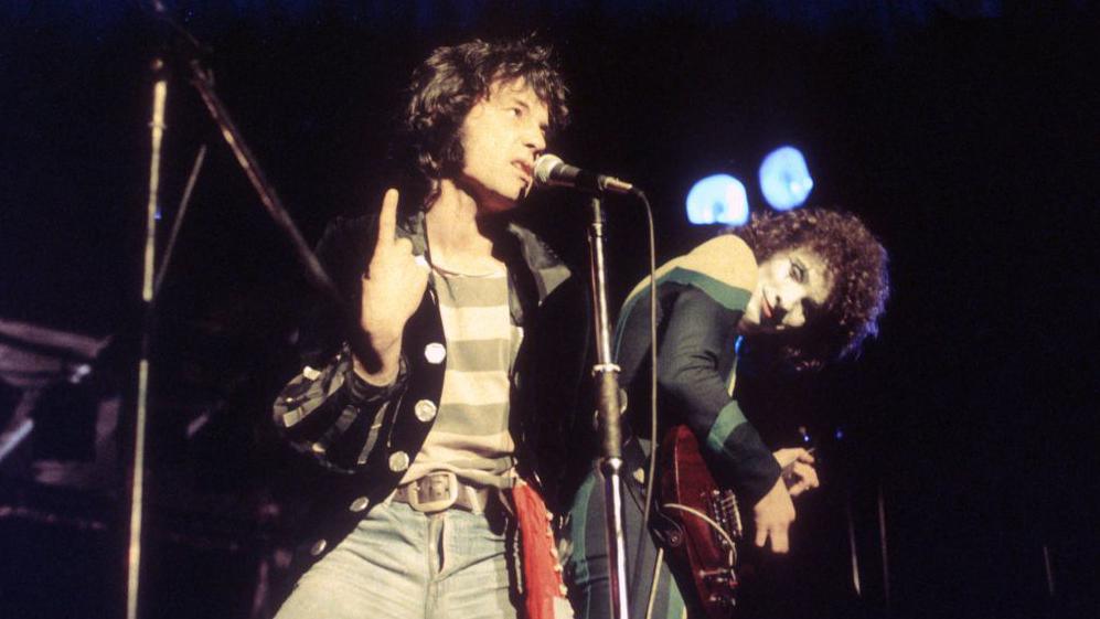 The Sensational Alex Harvey Band. Alex Harvey has dark, shoulder-length hair and is wearing a black jacket, a striped T-shirt and jeans. He is singing into a mic which is on a stand. A guitarist with dark, curly hair and face make-up is leaning over, looking at him