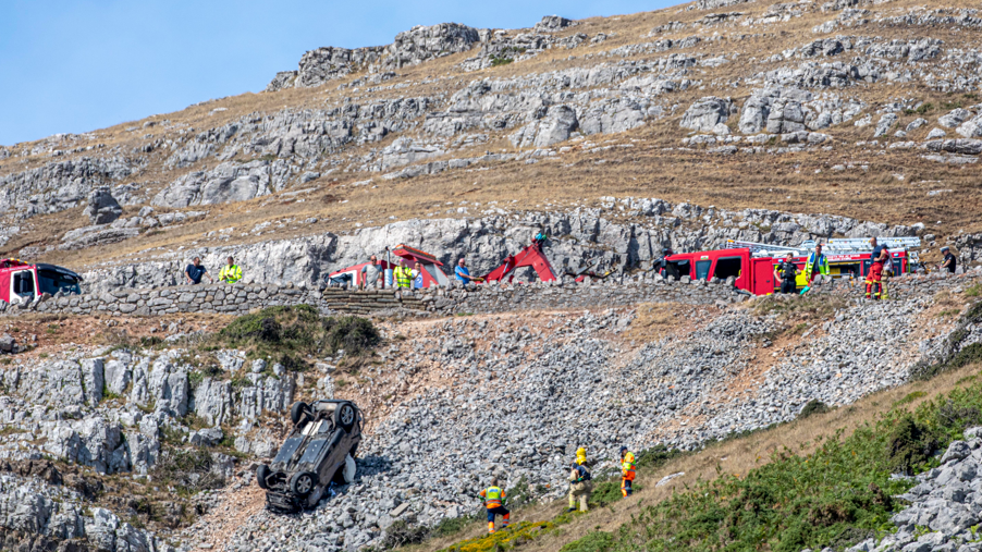 Upturned car on the side of a steep slope as emergency crews and vehicle recovery specialists work at the scene