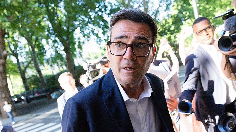Andy Burnham flanked by reporters and photographers on Millbank. He is wearing a white shirt with a black jacket and glasses. There are trees in the background.