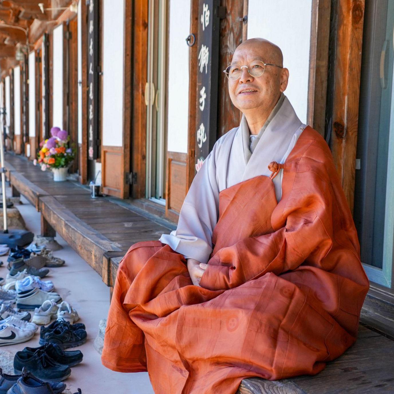 A Buddhist monk in orange and grey robes sits on the wooden porch of a traditional Korean temple building. Rows of shoes are lined up along the stone edge in front of the temple.