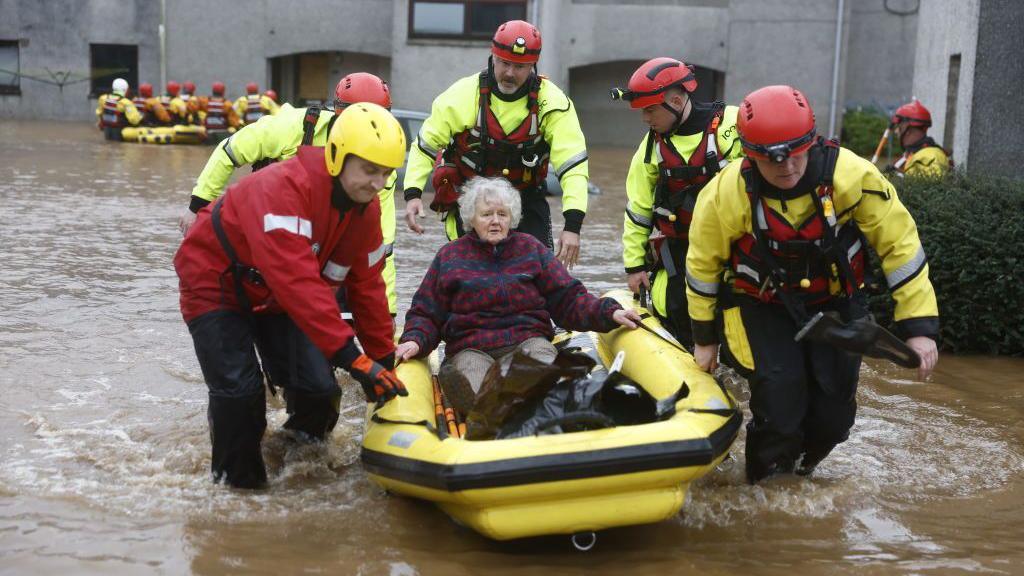 Coastguard volunteers carrying an older woman in a yellow raft in flood water in Brechin.