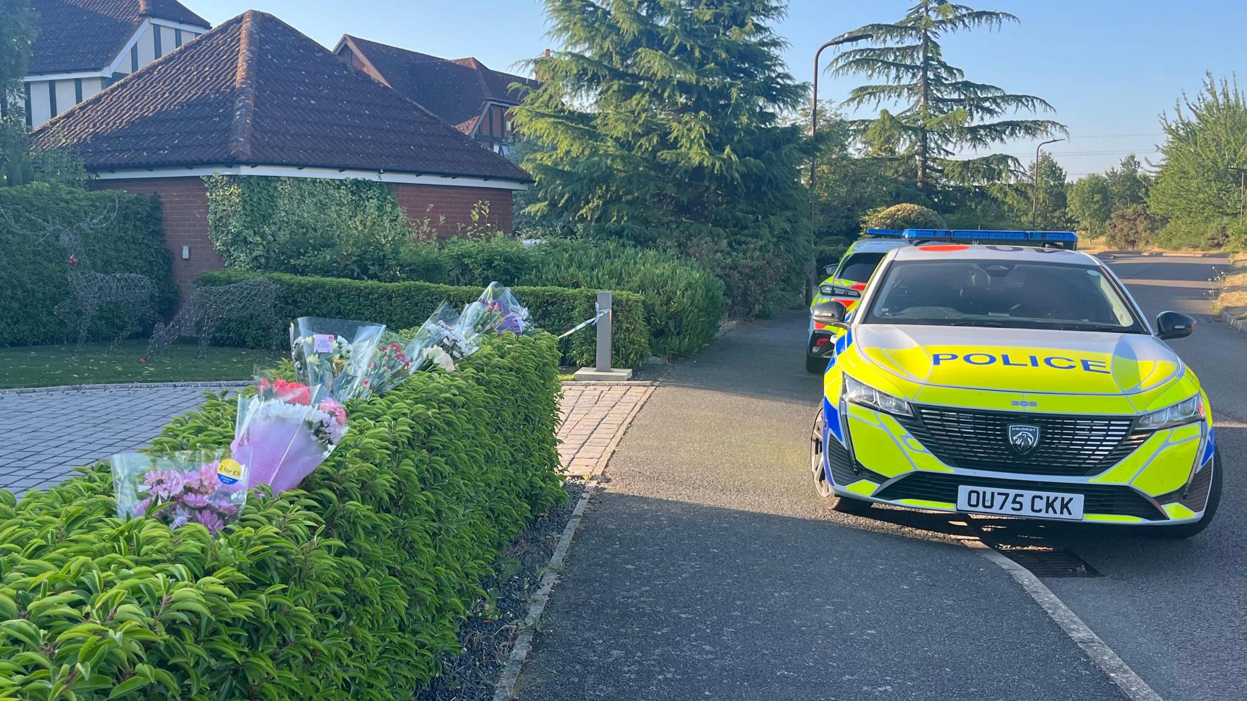 The scene at Carnoustie Drive in Great Denham. On the left is a hedge which has plastic-wrapped flowers sticking into it, and beyond is an entrance to a driveway with police tape across it. On the right are two police cars parked partly on the pavement. Beyond it is another detached house and trees.