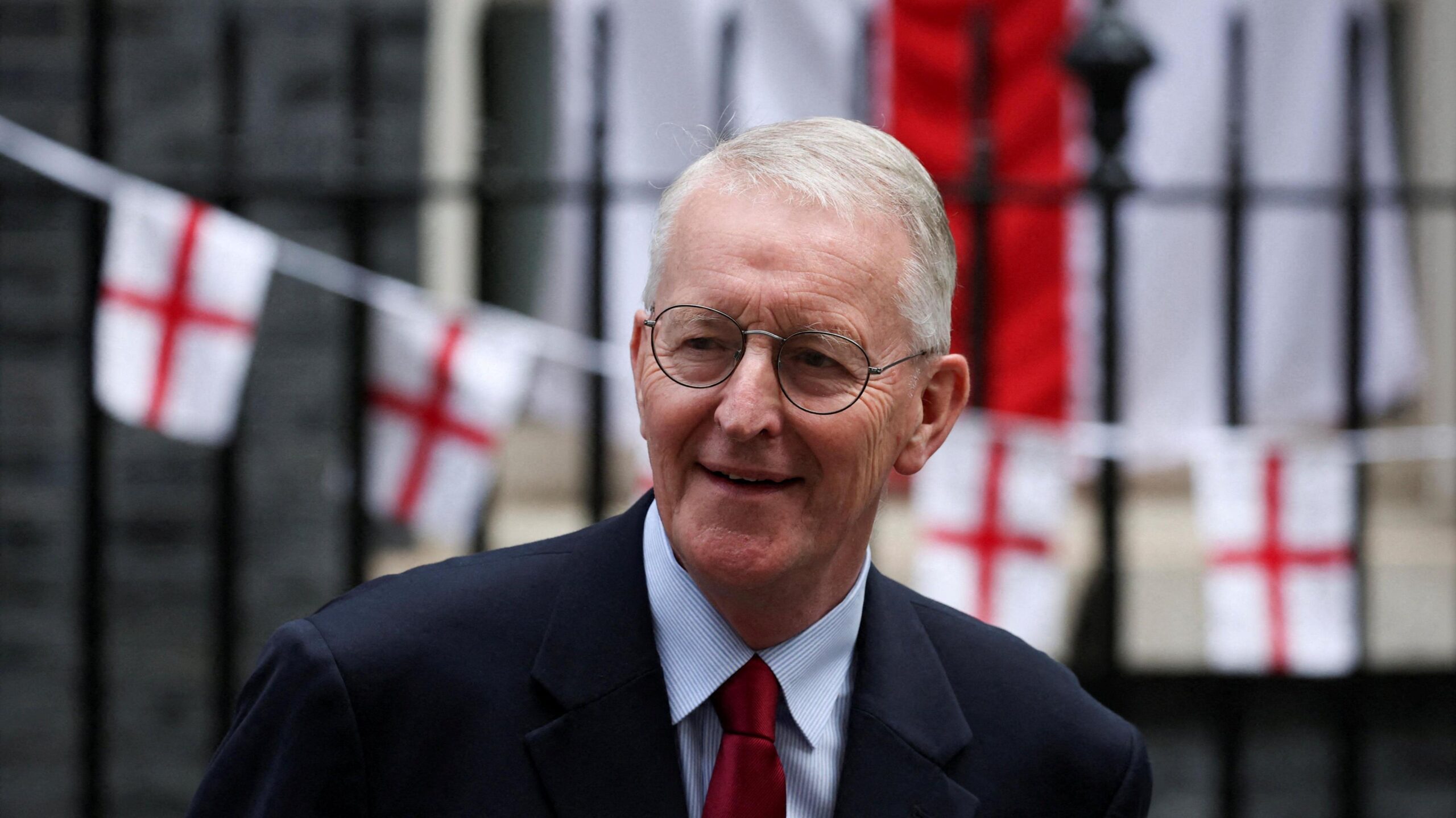 Hilary Benn, a man with short, white hair and metal-framed glasses, smiles as he walks out of 10 Downing Street. He is wearing a dark suit jacket, a light blue shirt and a red tie. In the background, bunting made of small red and white England flags is draped across black railings outside the prime minister's house.