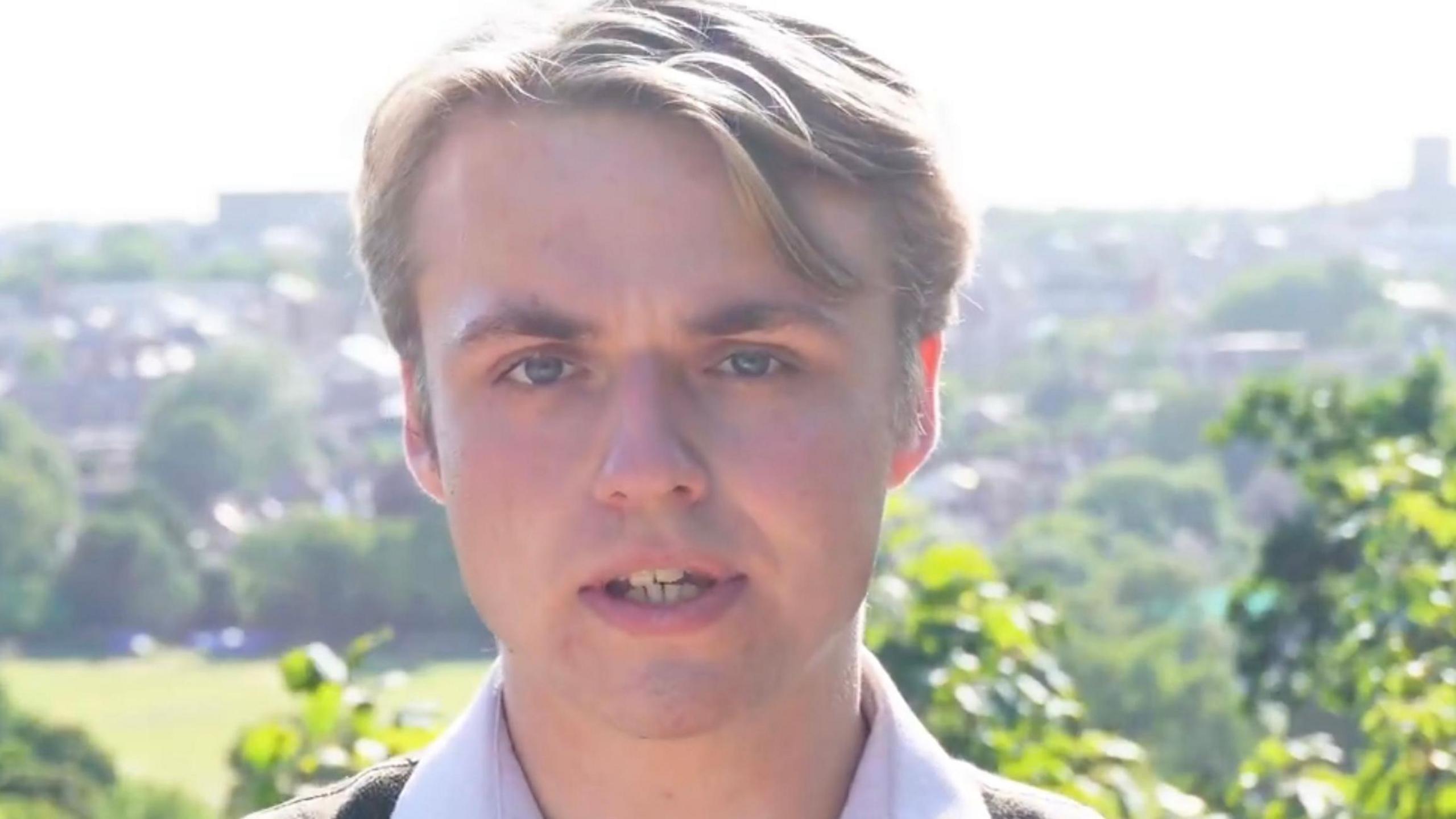 Kai Stephens with fair hair looks at the camera while delivering lines for a video recording. Behind him are trees and houses.