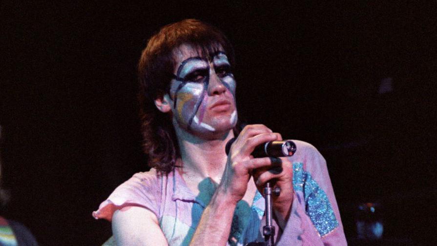 Fish from the band Marillion. He is on stage, holding a  microphone. His hair is in a mullet style and his face is covered in white and blue face paint. He is wearing a pink T-shirt with blue glitter on it and is glancing sideways at the camera.